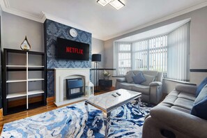 A living room with a fireplace, a sofa, a coffee table, and a TV mounted above the fireplace displaying the Netflix logo. - 3BR in Baxter Avenue With Netflix Wifi Backyard (Newcastle-upon-Tyne)