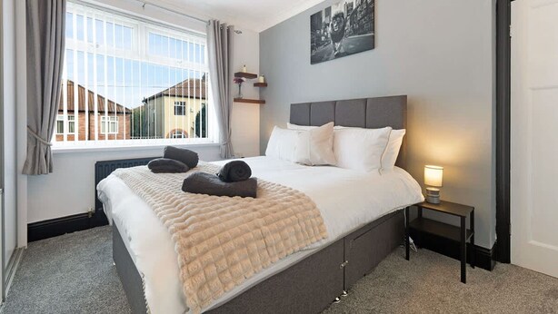 A hotel bedroom with a large bed, a bedside lamp, a window with curtains, and a wall-mounted picture. - 3BR in Baxter Avenue With Netflix Wifi Backyard (Newcastle-upon-Tyne)