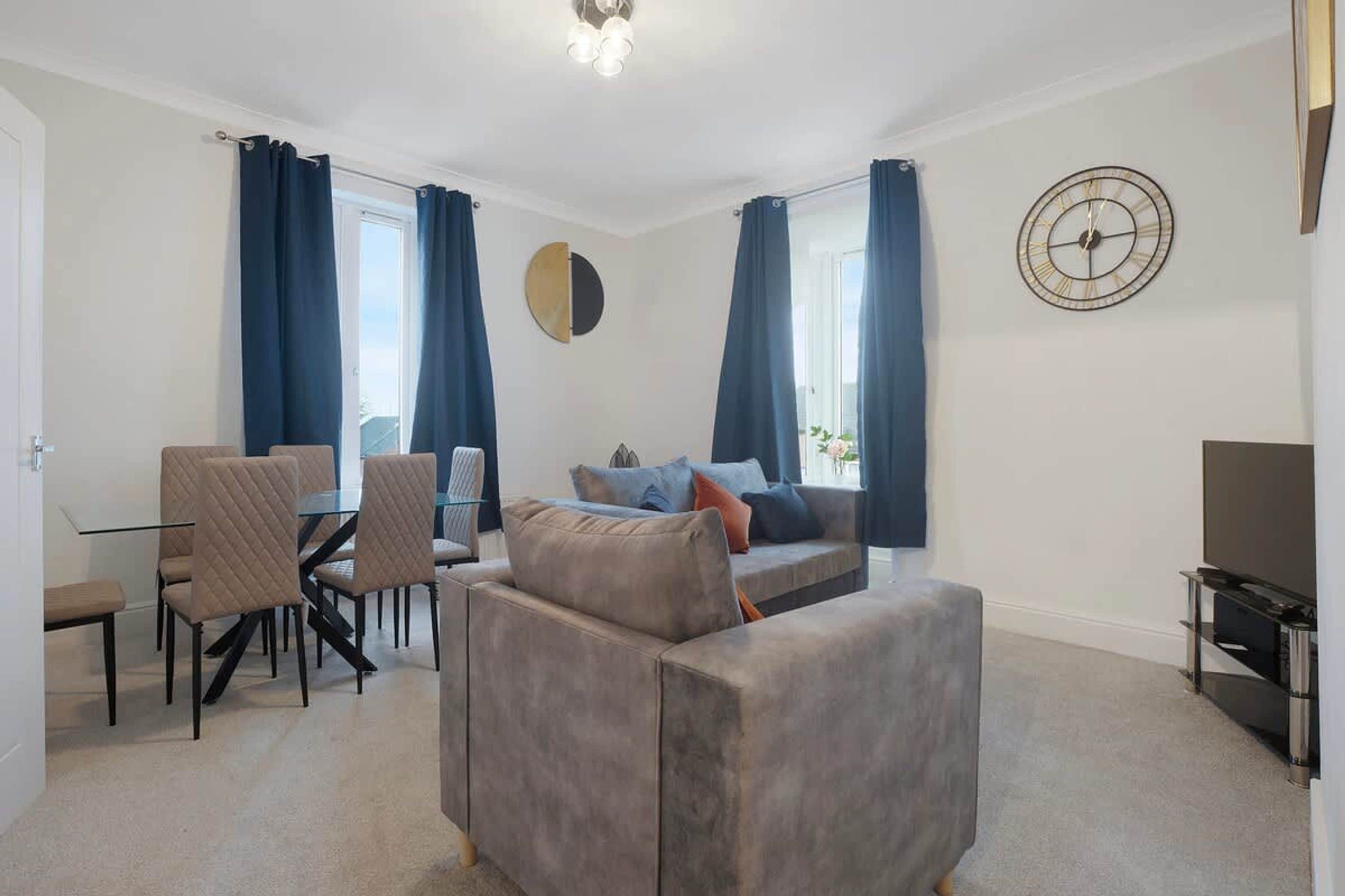 A modern living room with a sofa, a dining table with chairs, a TV, and a wall clock.