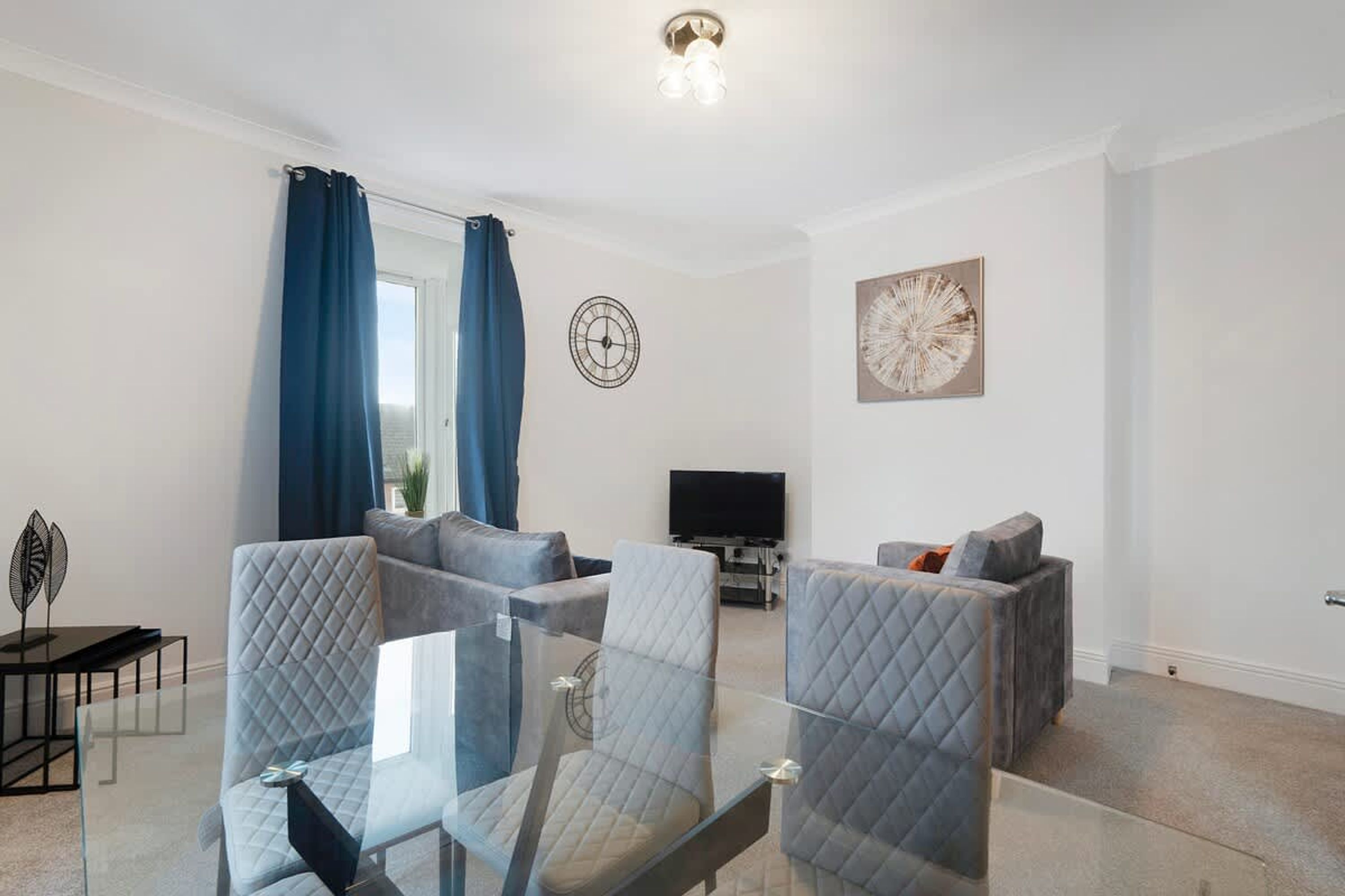 Un salon moderne avec une table basse en verre, deux fauteuils gris, un canapé, un téléviseur à écran plat et une horloge murale.