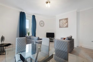A modern living room with a glass coffee table, two grey armchairs, a sofa, a flat-screen TV, and a wall clock. - Spacious 4 bed in North Shields With Free Wifi (North Shields)