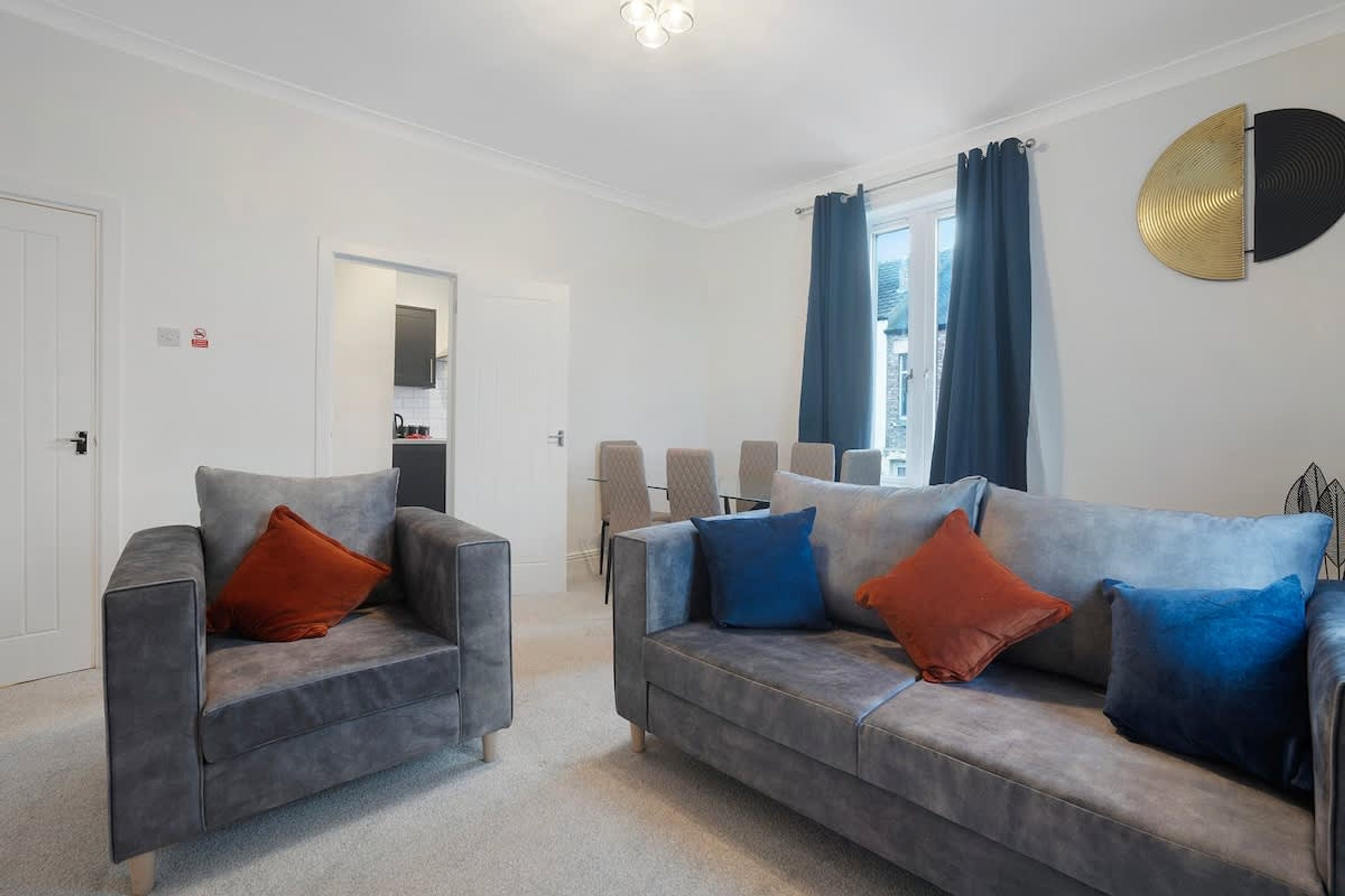 A living room with two grey sofas, one with orange and blue cushions, and a large window with blue curtains.