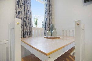 A small dining area with a wooden table, white chairs, a potted plant, and a window with curtains.