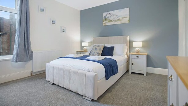 A bedroom with a large bed, bedside tables, a window with curtains, and a painting on the wall. - Seaside Haven on Newcomen Grove Redcar Retreat (Redcar)
