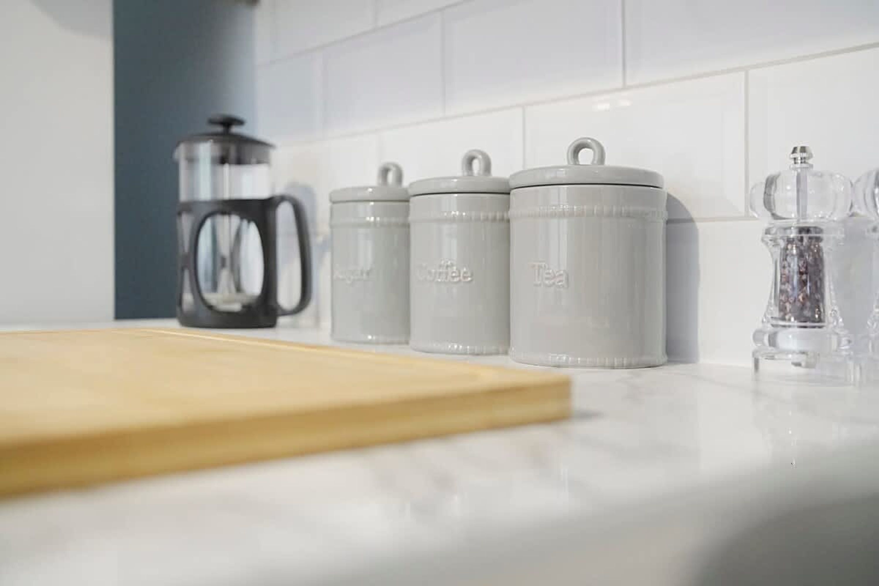 En køkkenbordplade med opbevaringskrukker mærket 'Sukker', 'Kaffe' og 'Te', en kaffemaskine og en klar glaskande.