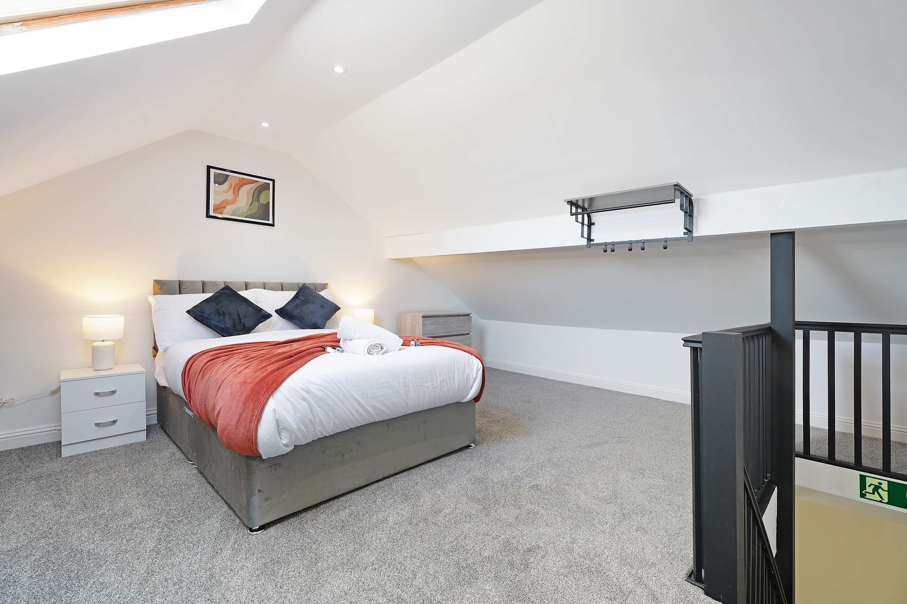Une chambre à coucher comprenant un lit, des tables de chevet, une commode, un luminaire au plafond, une étagère fixée au mur et une rambarde noire.