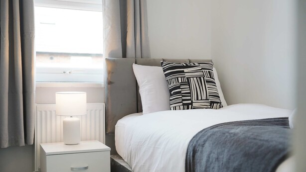 A neatly made bed with a white comforter and a black and white patterned pillow, a white bedside table with a lamp, and grey curtains on a window. - Comfortable 4BR Stay Byerley Road by Stay With Us (Shildon)