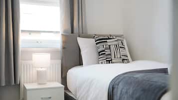 A neatly made bed with a white comforter and a black and white patterned pillow, a white bedside table with a lamp, and grey curtains on a window.