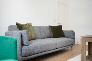 A grey sofa with two cushions, a wooden side table, and a green chair in a room with white walls and a wooden floor. - Comfortable 4BR Stay Byerley Road by Stay With Us (Shildon)