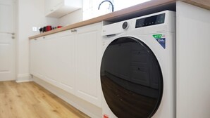 A front view of a modern, white washing machine with a digital control panel, located in a room with wooden flooring and white cabinetry. - Comfortable 4BR Stay Byerley Road by Stay With Us (Shildon)