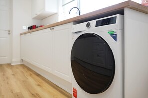 A front view of a modern, white washing machine with a digital control panel, located in a room with wooden flooring and white cabinetry. - Comfortable 4BR Stay Byerley Road by Stay With Us (Shildon)