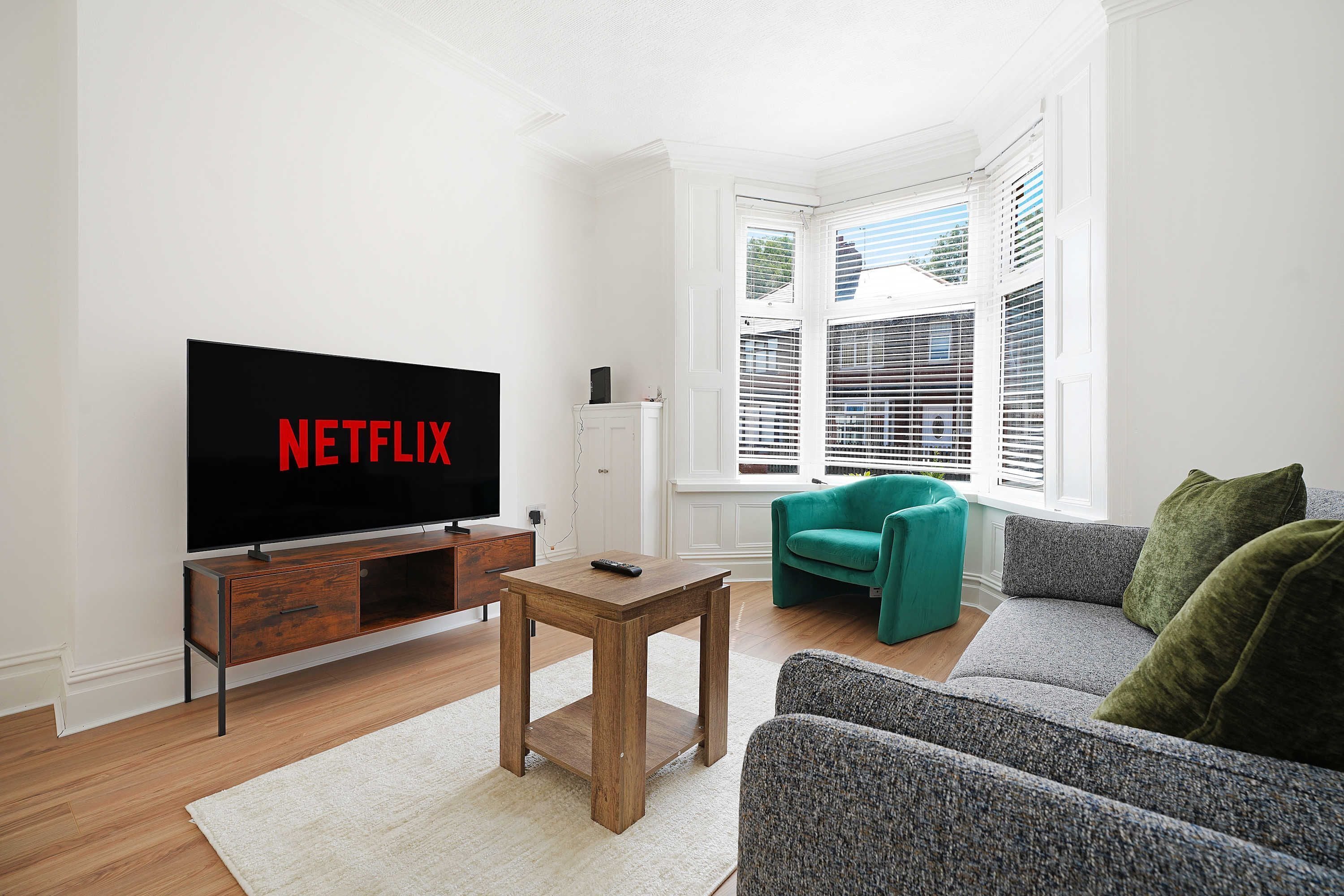 A living room with a television displaying 'NETFLIX,' a wooden coffee table, a teal armchair, and a grey sofa.