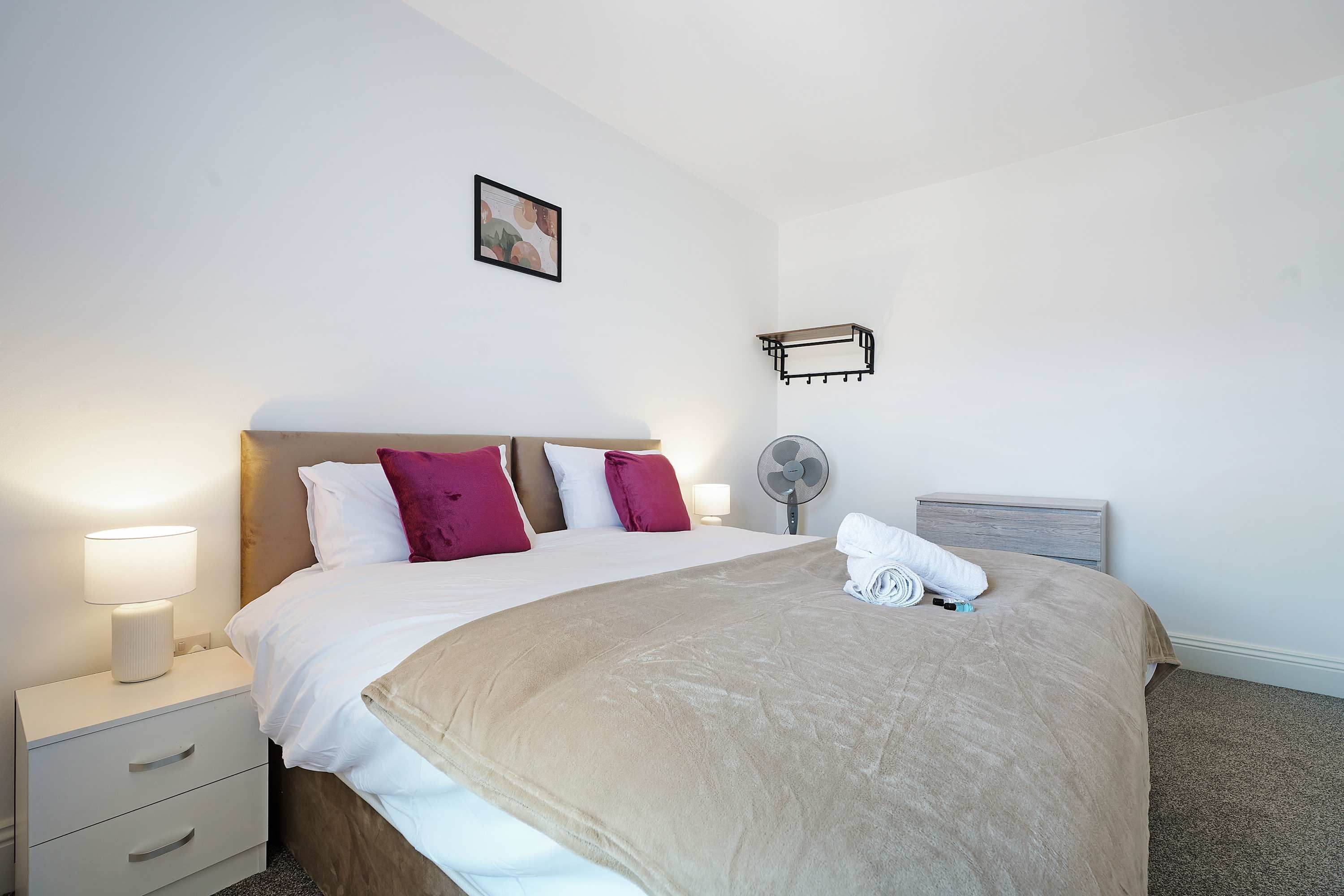 A neatly made bed with a beige blanket, white and maroon pillows, a small bedside table with a lamp, a wall-mounted fan, and a picture frame on the wall.