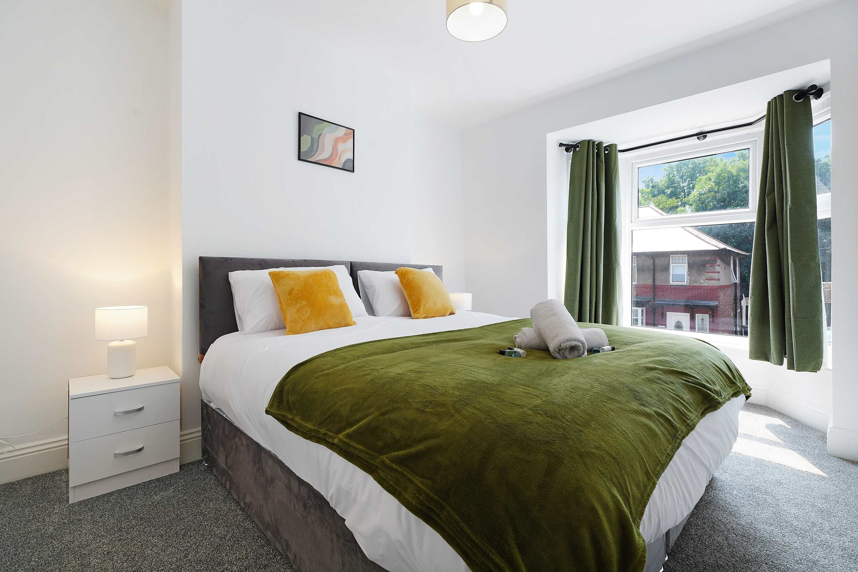 A hotel room with a bed, a bedside table with a lamp, a window with curtains, and a view of the outside.