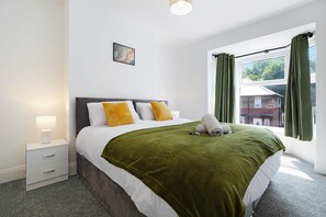 A hotel room with a bed, a bedside table with a lamp, a window with curtains, and a view of the outside. - Comfortable 4BR Stay Byerley Road by Stay With Us (Shildon)