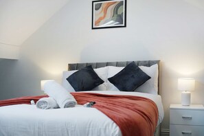 Una cama bien hecha con almohadas y una colcha, una mesita de noche con una lámpara, y un cuadro abstracto enmarcado en la pared.