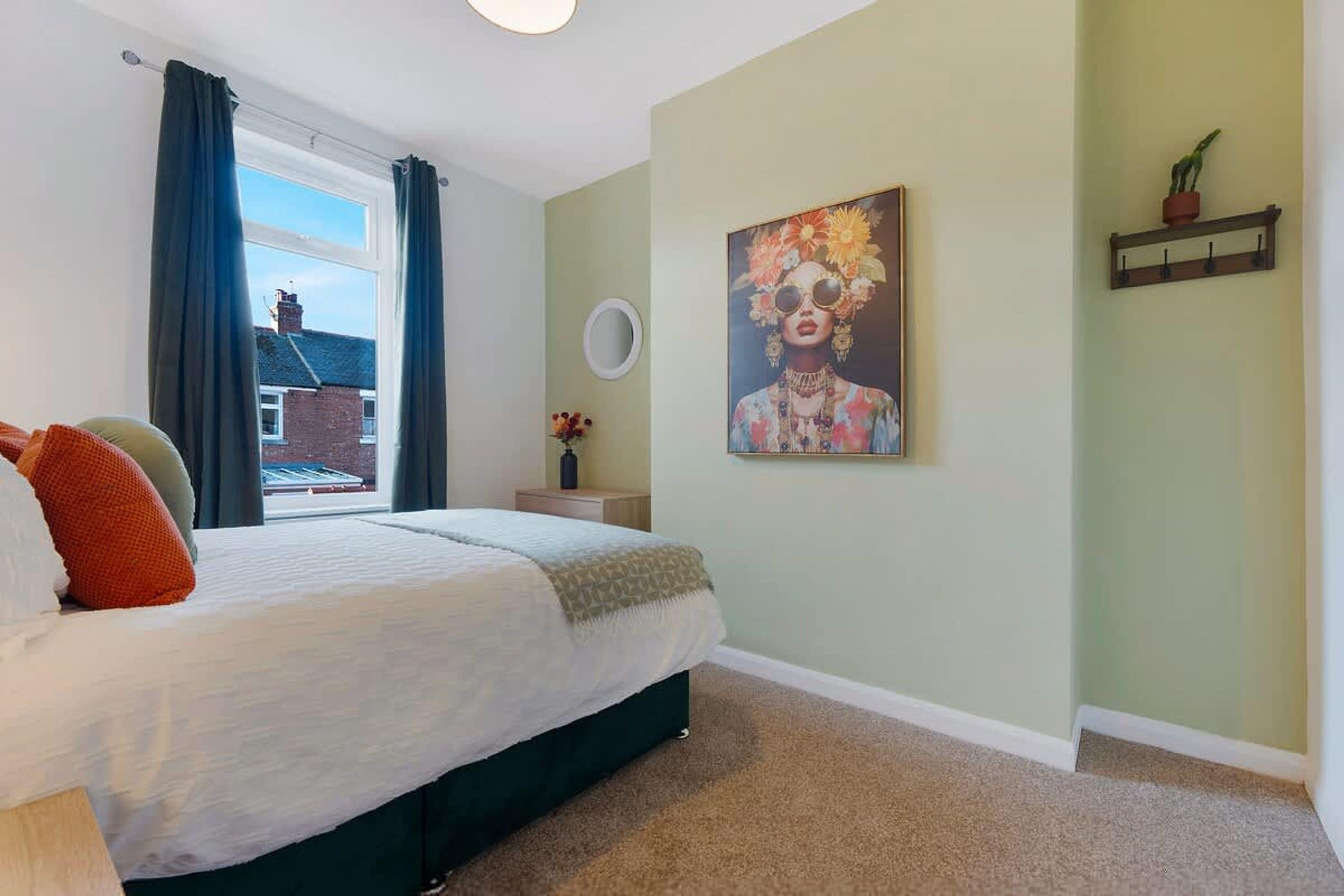 A bedroom with a bed, a window with curtains, a painting on the wall, and a small plant on a shelf.