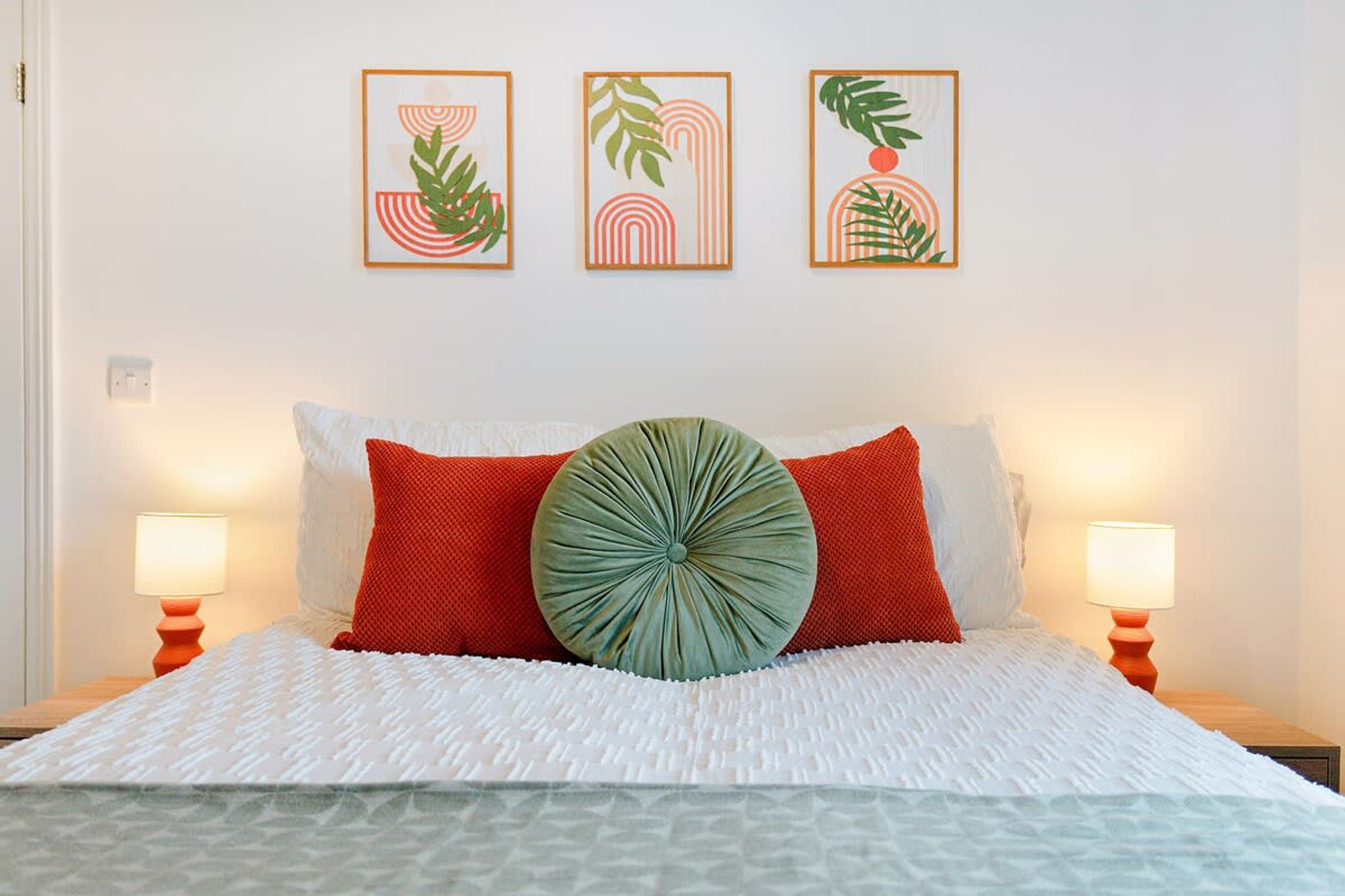 A bed with a white bedspread, red and green pillows, and a round green accent pillow. There are three framed artworks above the bed.