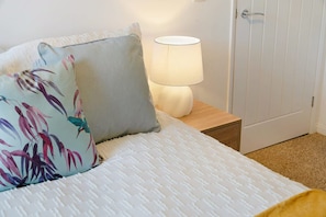 Una cama bien hecha con una colcha blanca con textura, dos almohadas estampadas con motivos florales, una lámpara de noche y una mesita de madera.