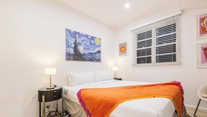 Un dormitorio con una cama, dos mesitas de noche con lámparas, una ventana con persianas y un cuadro en la pared.