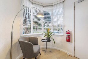 Habitación de esquina con una silla, una mesita con planta en maceta, una lámpara de pie y un extintor de incendios colgado en la pared.
