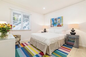Un dormitorio con cama, mesita de noche, lámpara, ventana con persianas, un colorido tapete a rayas y un cuadro en la pared.