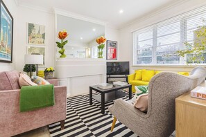 Ein modernes Wohnzimmer mit Sofa, Sessel und Couchtisch. Ein Fernseher und ein Fenster mit Jalousien sind ebenfalls vorhanden.