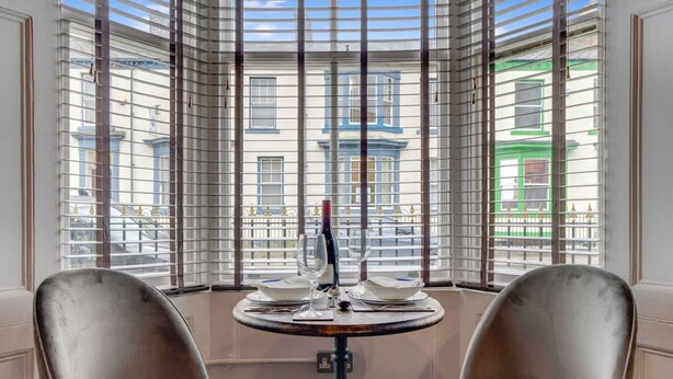 Angolo pranzo con tavolo rotondo e due sedie, una finestra con persiane e vista sugli edifici esterni.