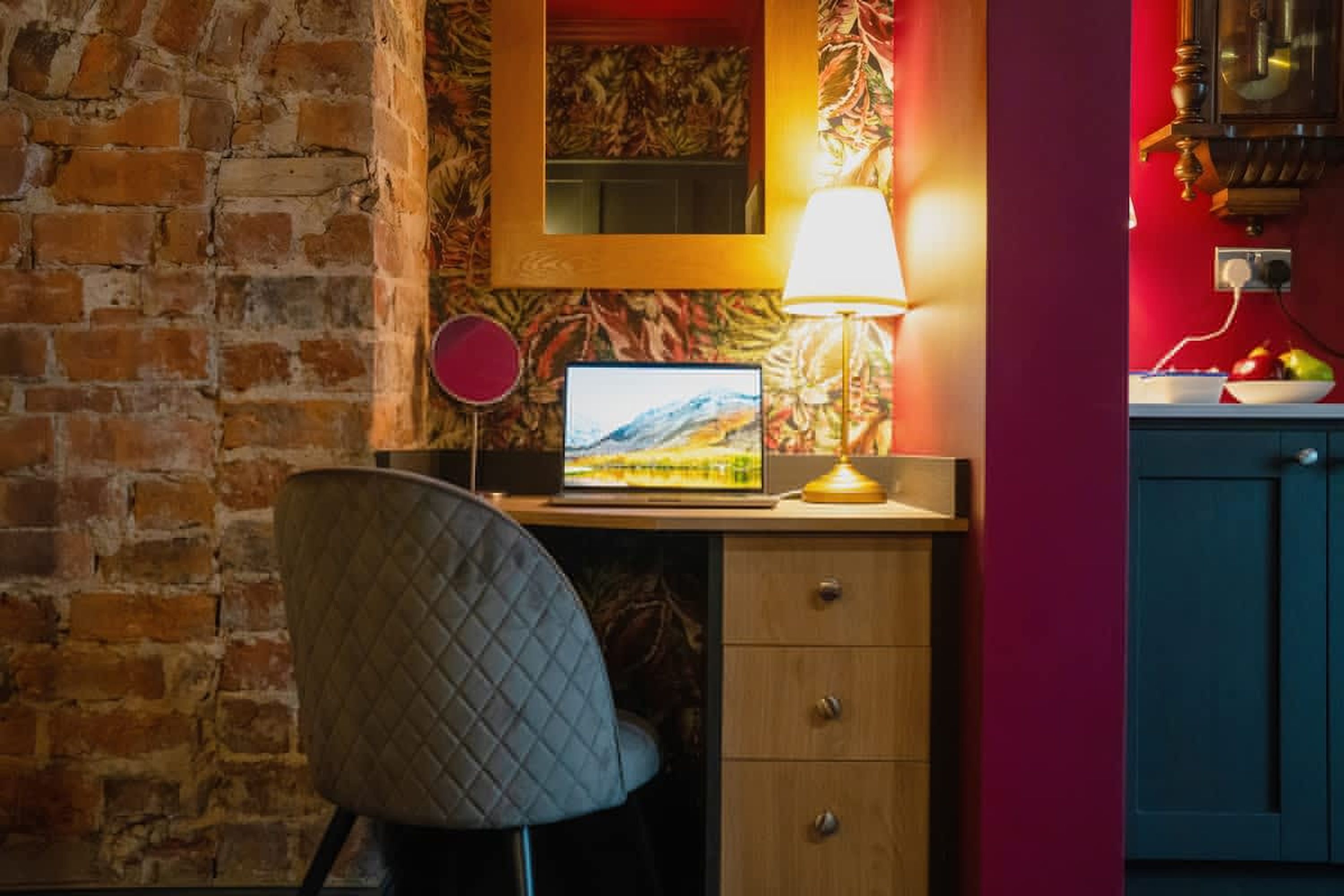 A room with a brick wall, a desk with a laptop, a chair, a lamp, and a red wall with a painting.