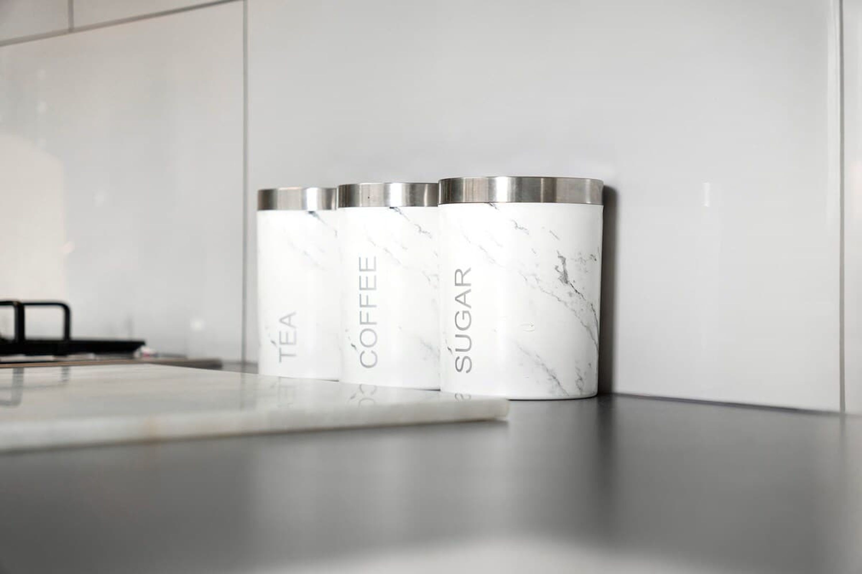 Three white marble containers with metallic tops labeled 'TEA,' 'COFFEE,' and 'SUGAR' on a kitchen counter.