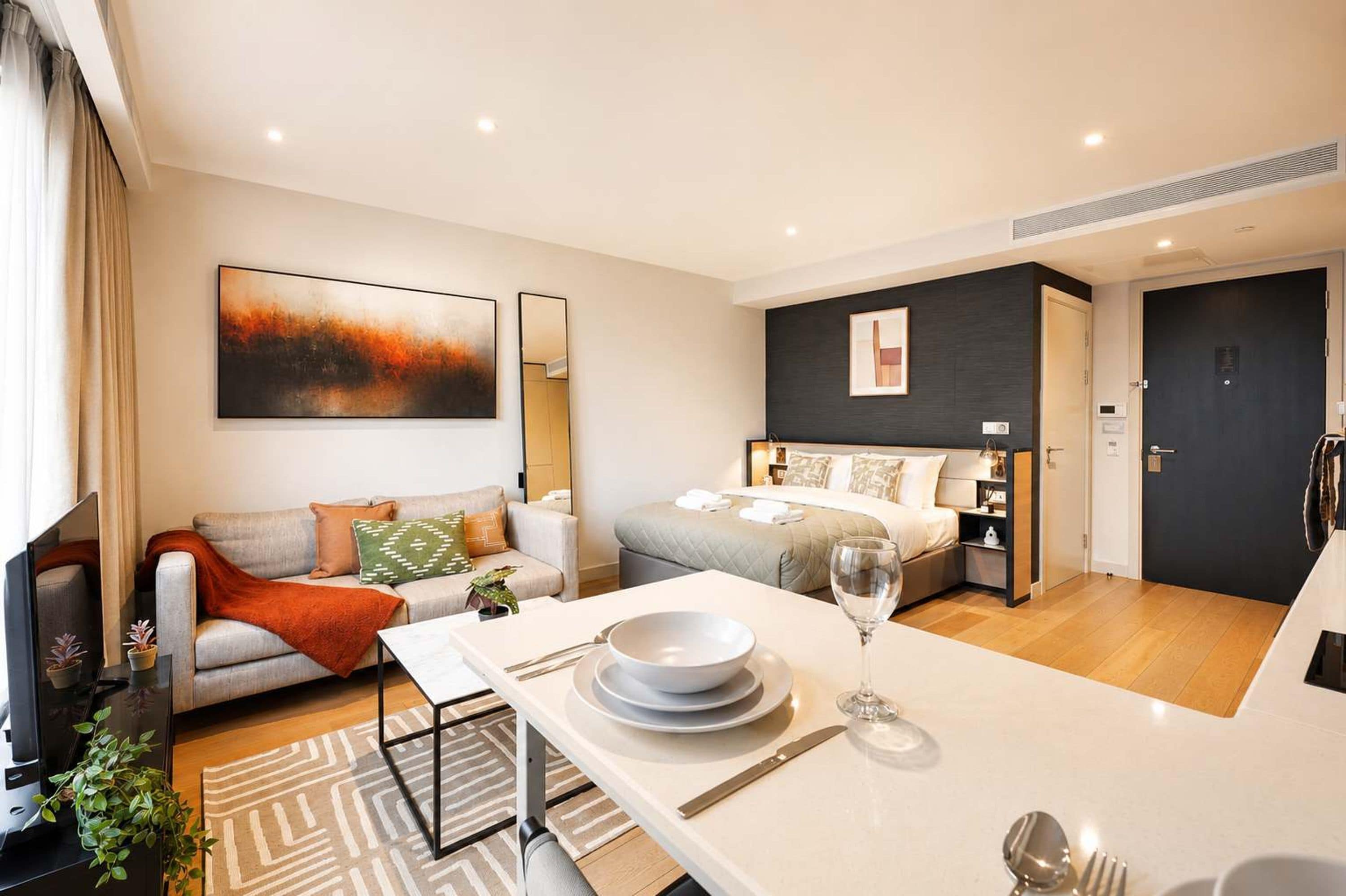 Habitación de hotel moderna con cama, sofá, una mesa de comedor con platos y cubiertos, y un cuadro abstracto grande en la pared.