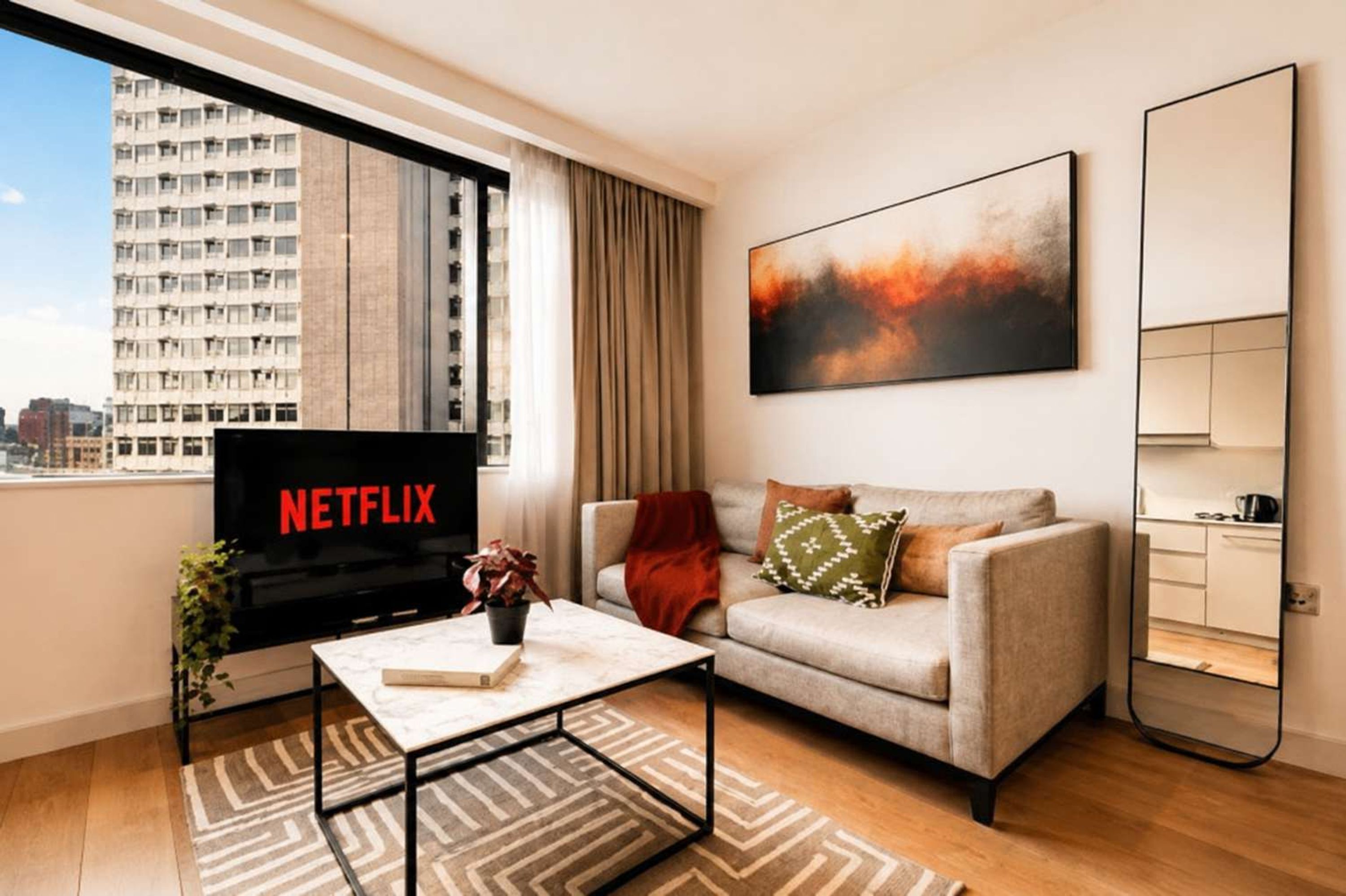 A modern living room with a sofa, a coffee table, a TV displaying 'NETFLIX', and a city view through the window.