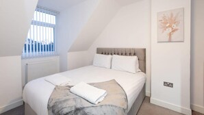 Une chambre avec un plafond en pente, un lit avec du linge de lit blanc, un radiateur, une fenêtre avec des stores et un interrupteur fixé au mur.