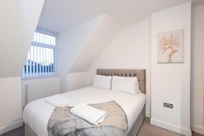 A bedroom with a sloped ceiling, a bed with white linens, a radiator, a window with blinds, and a wall-mounted light switch. - Beautiful two Bedroom Family Home With Pool Table Durham (Durham)