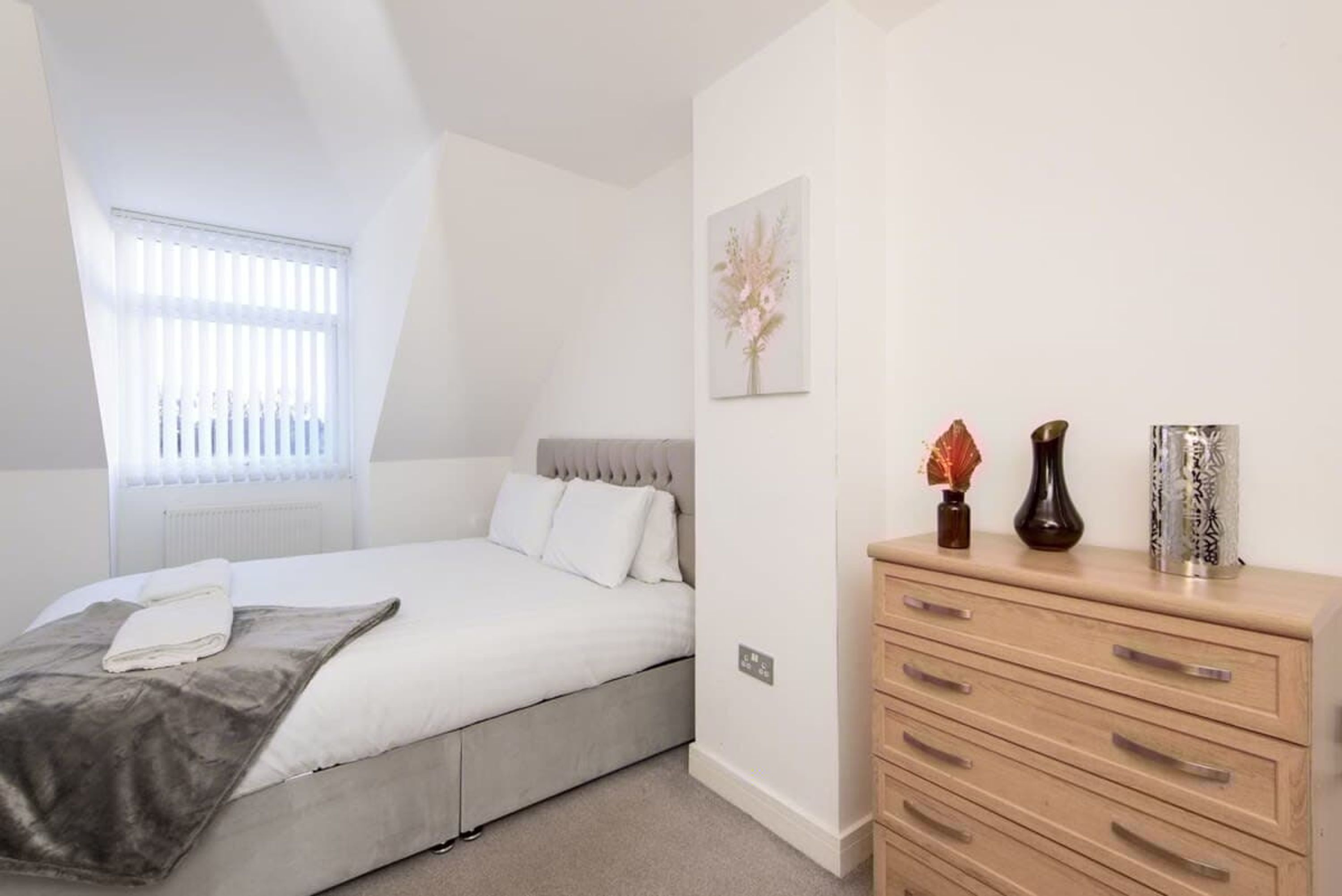 A bedroom with a bed, a dresser, a window with blinds, and a wall-mounted light switch.