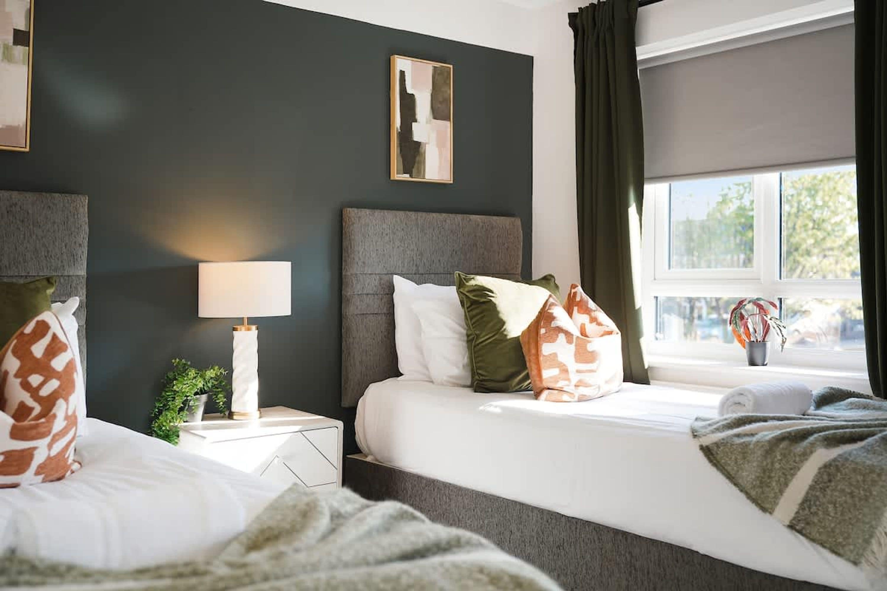 A hotel room with two beds, a nightstand, a lamp, a window with curtains, and a framed picture on the wall.