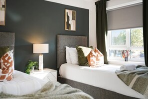 A hotel room with two beds, a nightstand, a lamp, a window with curtains, and a framed picture on the wall. - Modern Coastal Stay Minutes From Redcar Beach (Redcar)