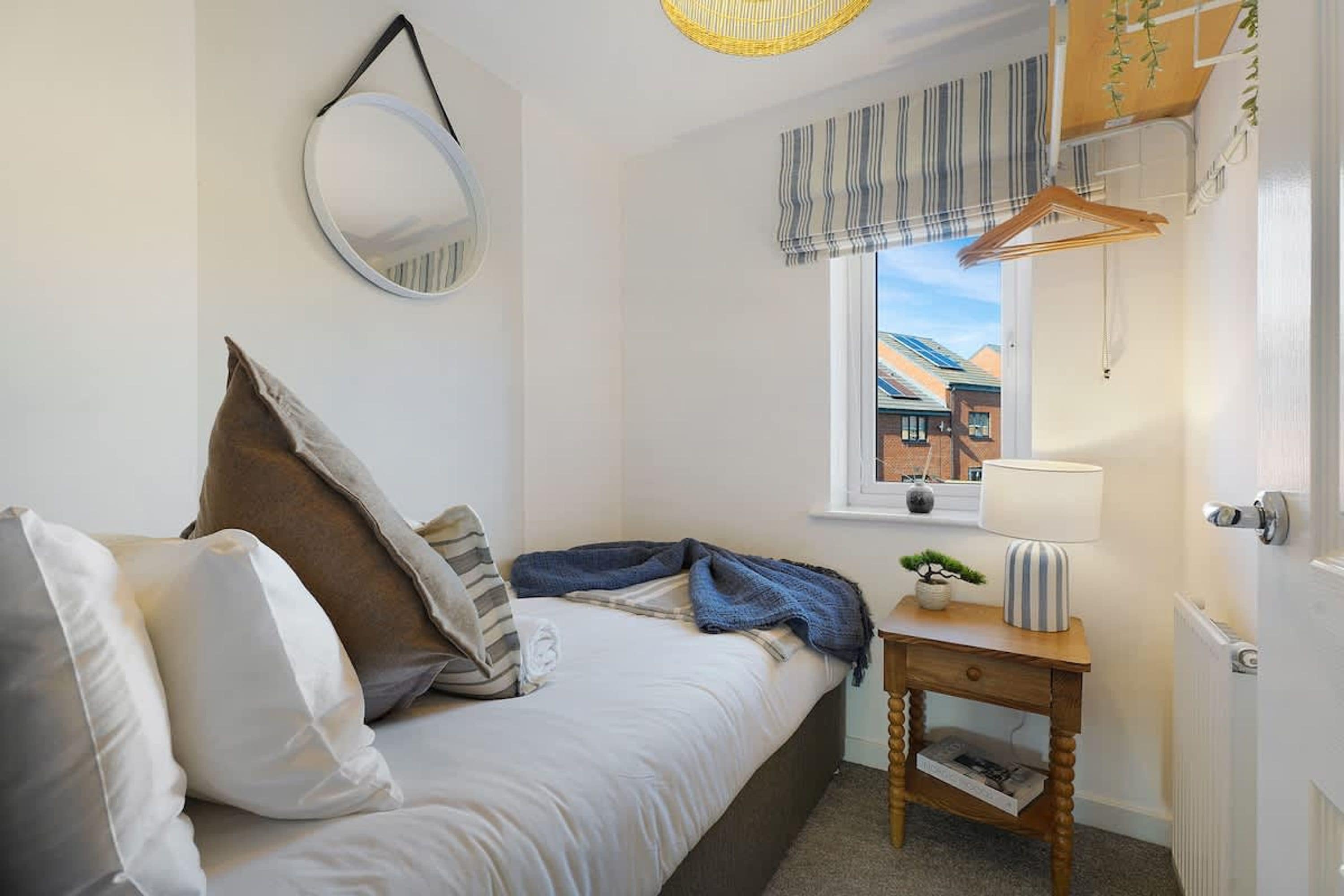 A bedroom with a bed, pillows, a bedside table with a lamp, a hanging mirror, and a window with a view of buildings.