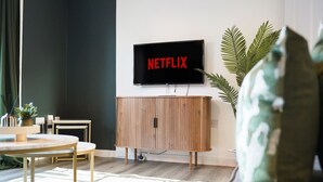 Olohuoneessa on seinälle asennettu televisio, jossa näkyy teksti ’NETFLIX’. Sen alla on puinen kaappi, ja huoneessa on pyöreä pöytä tuoleineen sekä suuri viherkasvi.