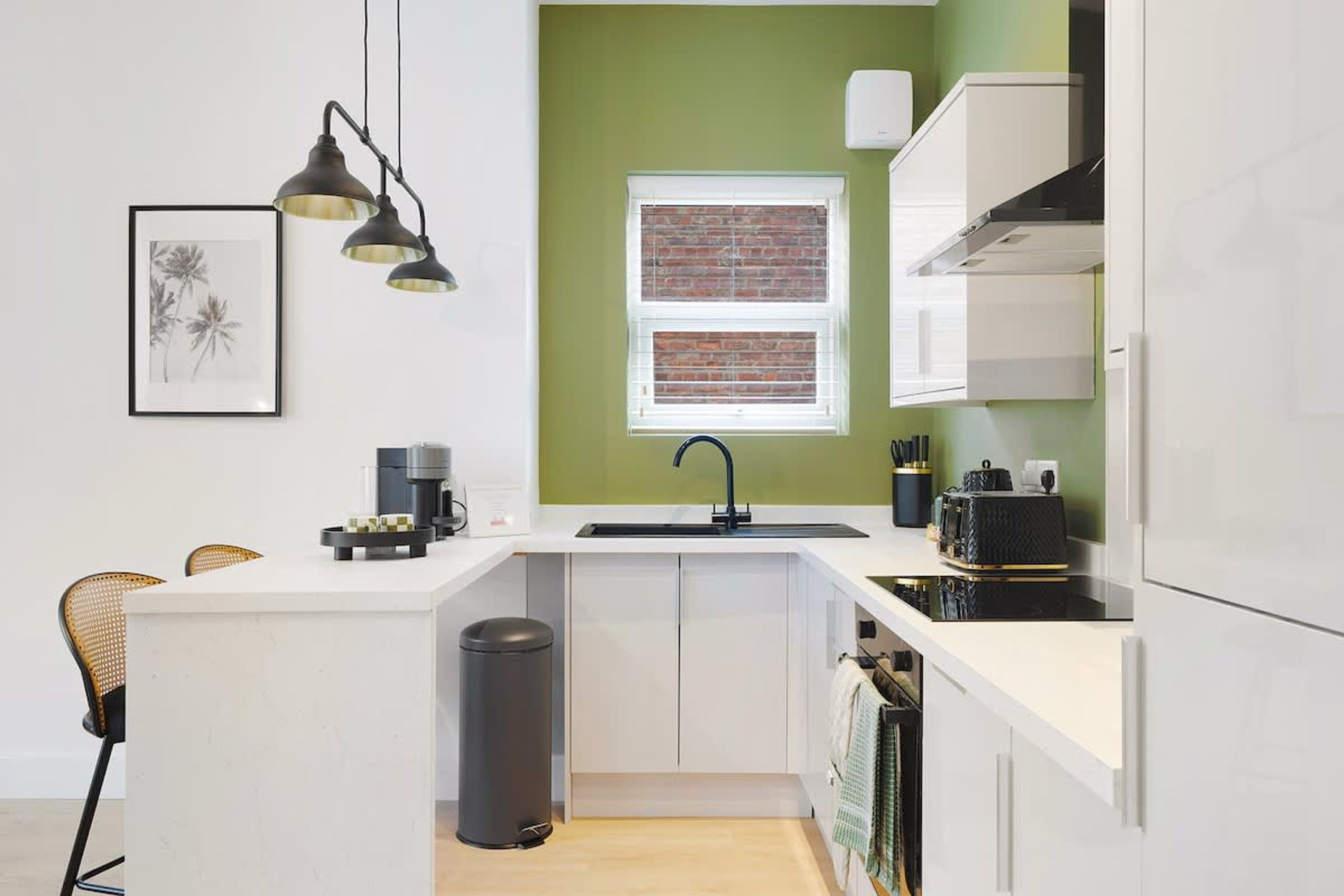 Una cucina moderna con mobili bianchi, un cestino nero, una macchina per il caffè e una finestra con vista su un muro di mattoni.
