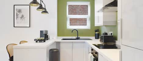 Una cucina moderna con mobili bianchi, un cestino nero, una macchina per il caffè e una finestra con vista su un muro di mattoni.