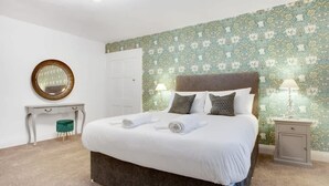 Ein ordentlich bezogenes Bett mit weißen Leinen und zwei Kissen, ein kleiner Nachttisch mit Lampe, ein runder Spiegel an der Wand und ein grüner Hocker.