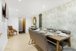 Un coin repas avec une longue table dressée pour six personnes, un téléviseur fixé au mur et une porte donnant sur une autre pièce.