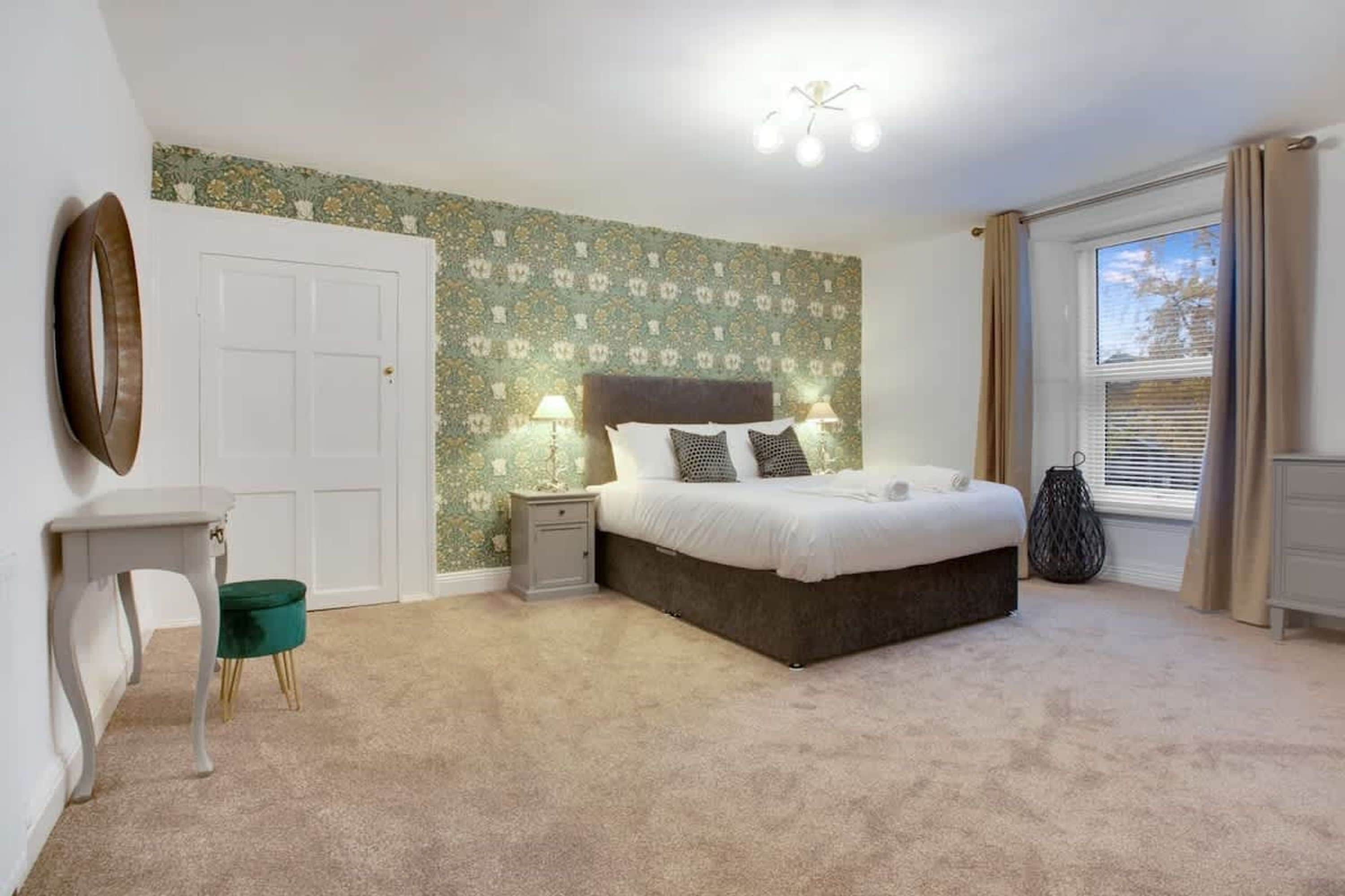 A bedroom with a bed, bedside tables, a chair, a window with curtains, and a wall with a floral pattern.
