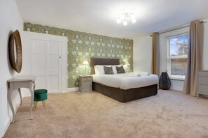 Un dormitorio con cama, mesitas de noche, una silla, una ventana con cortinas y un papel tapiz con motivos florales.