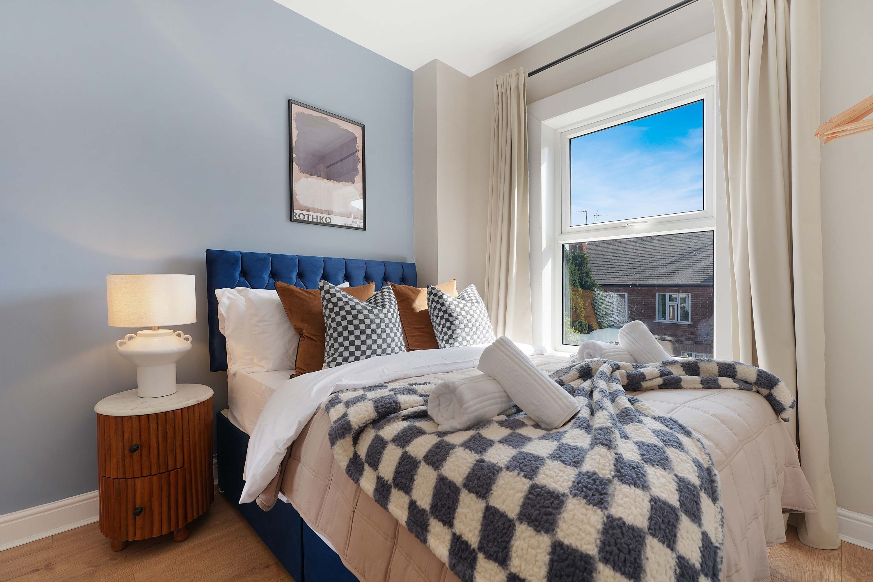 Une chambre à coucher comprenant un lit, des oreillers, une table de chevet, une lampe, une fenêtre avec des rideaux et un tableau accroché au mur.