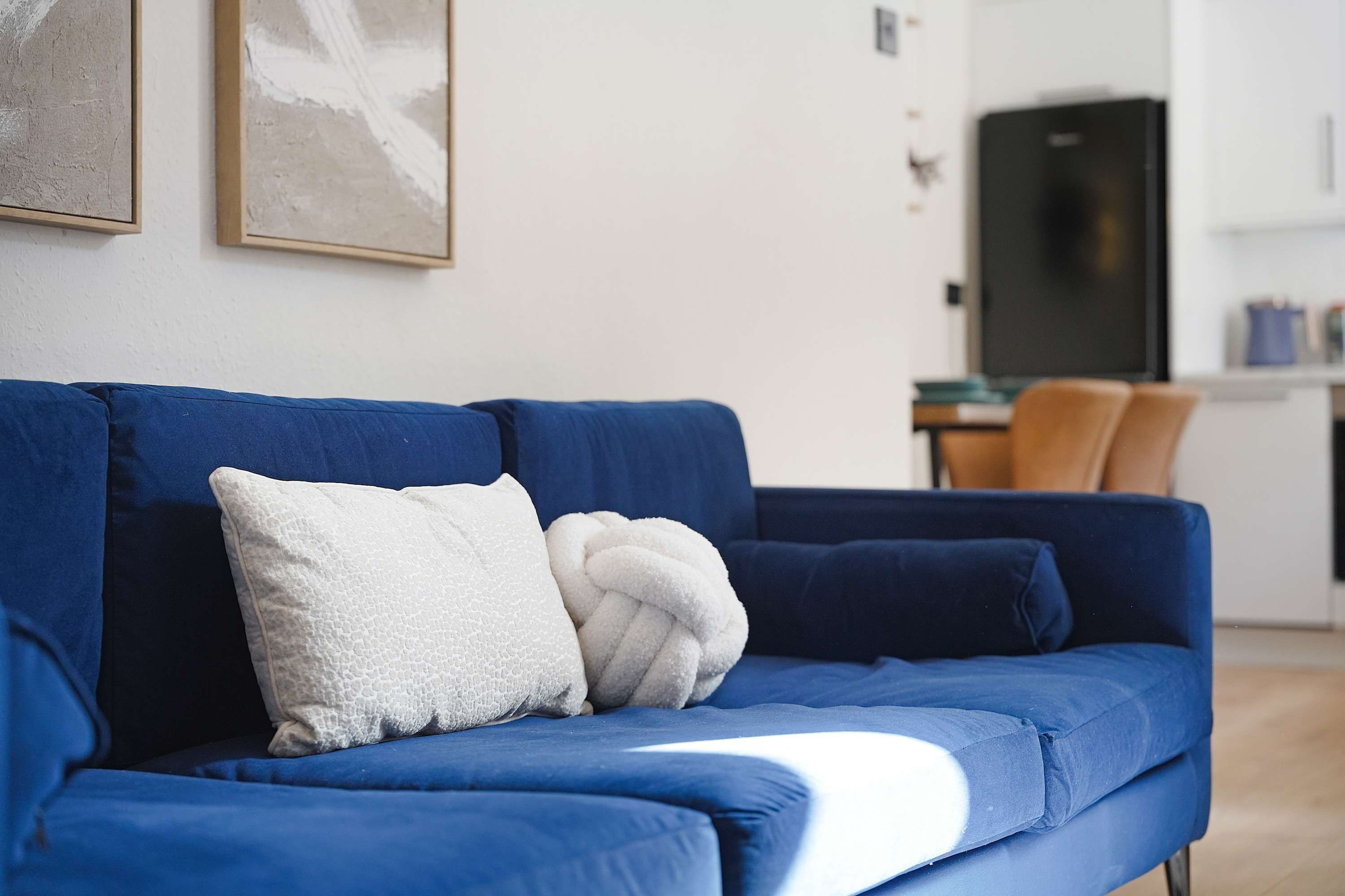 A blue sofa with a white pillow and a white throw blanket in a modern living room with a kitchen in the background.