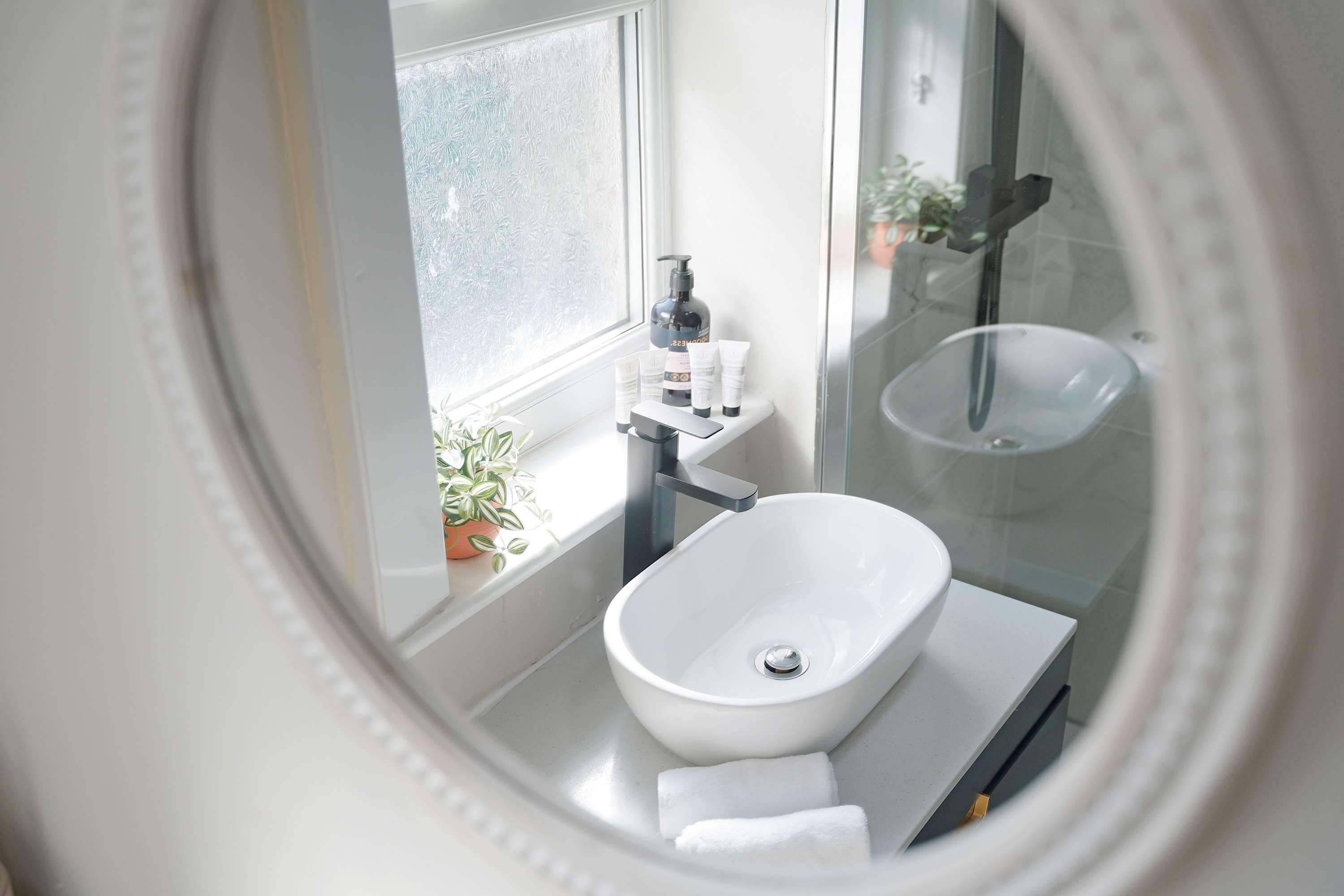 Een badkamer met een wit wastafelmeubel, een glazen douchecabine, een raam met geëtste ruit en een potplant op de vensterbank.