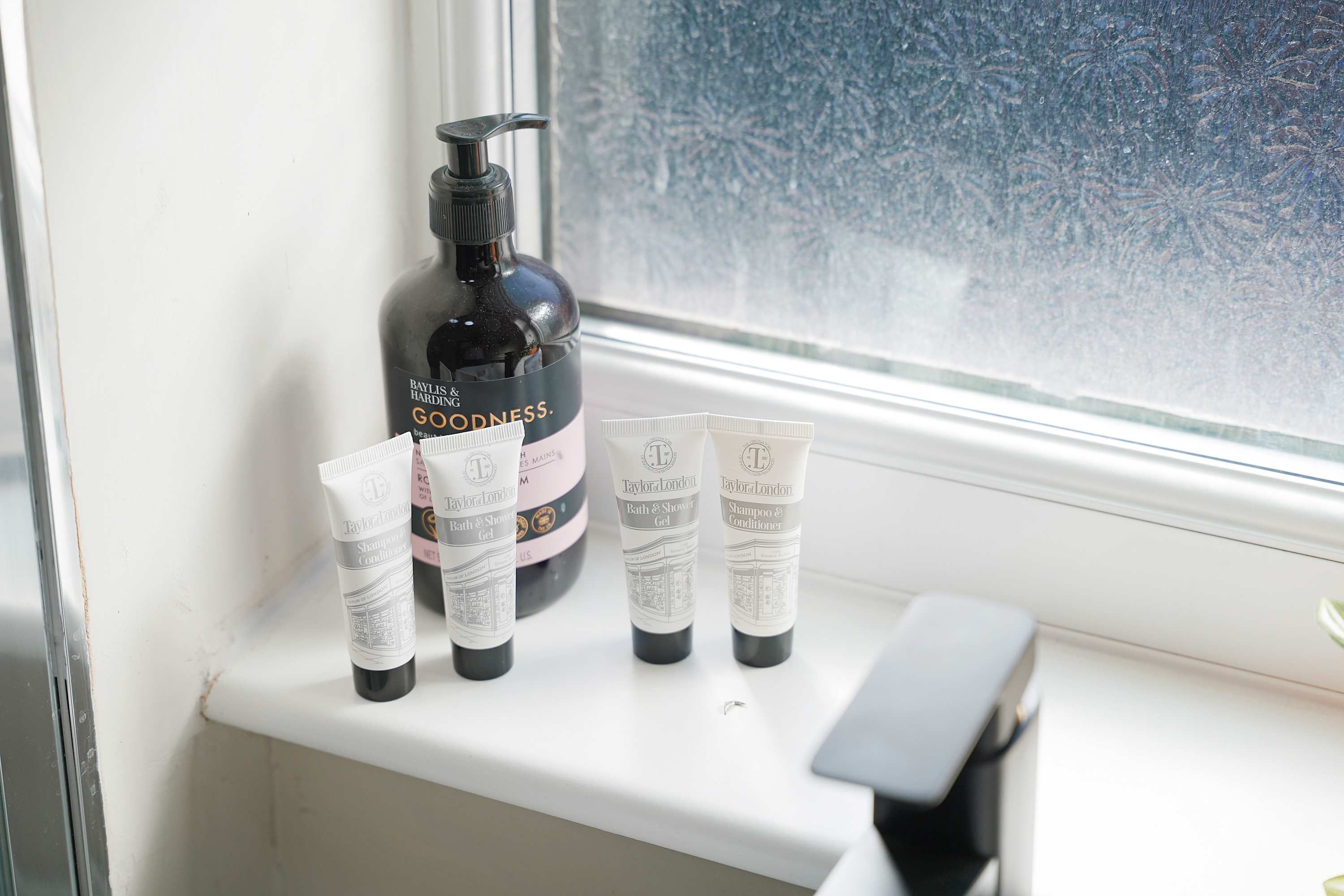 Ein Badezimmerschrank mit einer Flasche Baylis & Harding Goodness, vier Tuben Taylor of London Bath & Shower Gel und einem schwarzen Gegenstand.