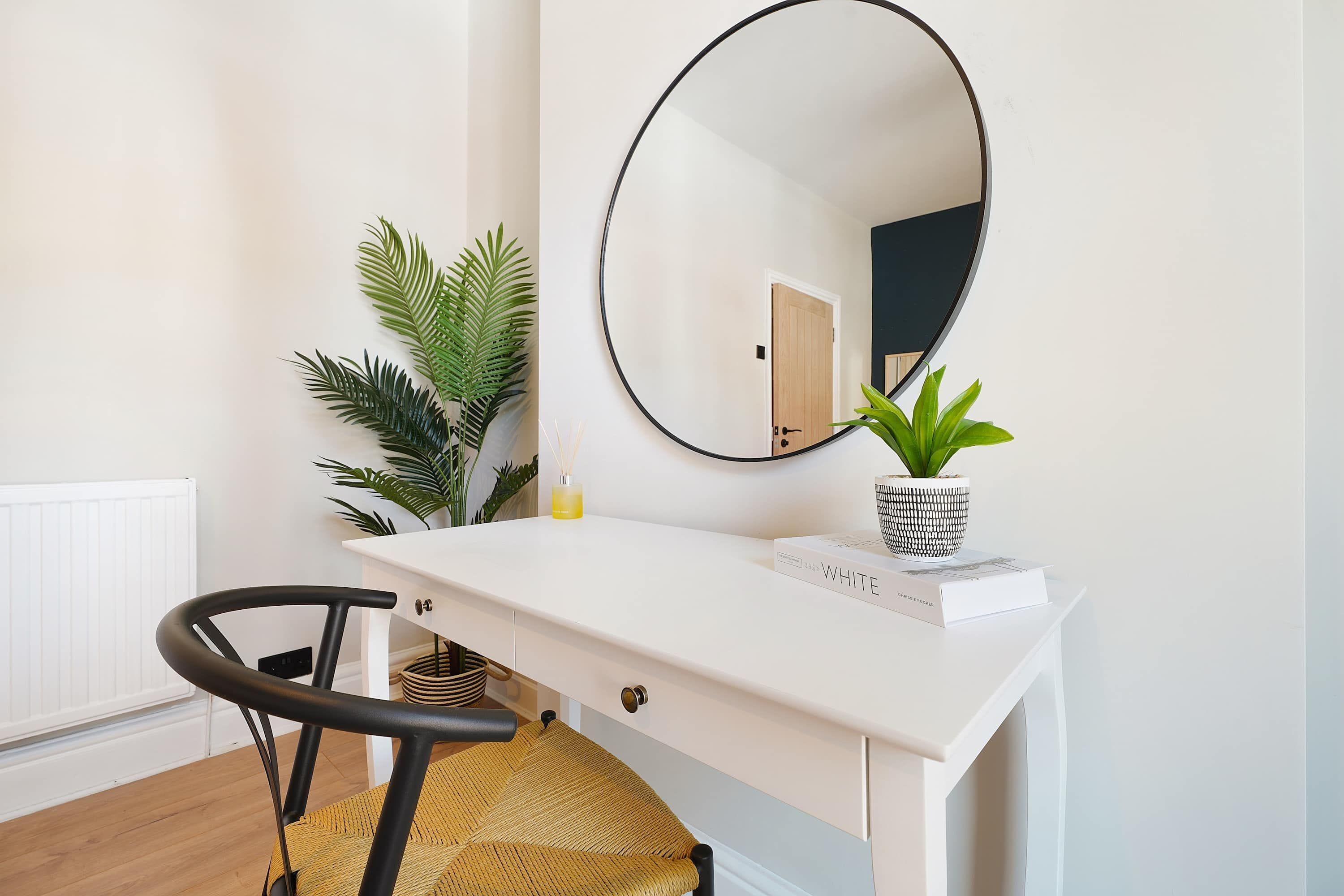 Un bureau blanc avec un miroir rond, une plante en pot, un livre et une chaise.